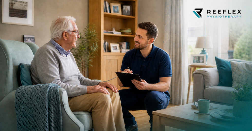 Older man seated in an armchair at home talking through his rehabilitation plan with a physiotherapist